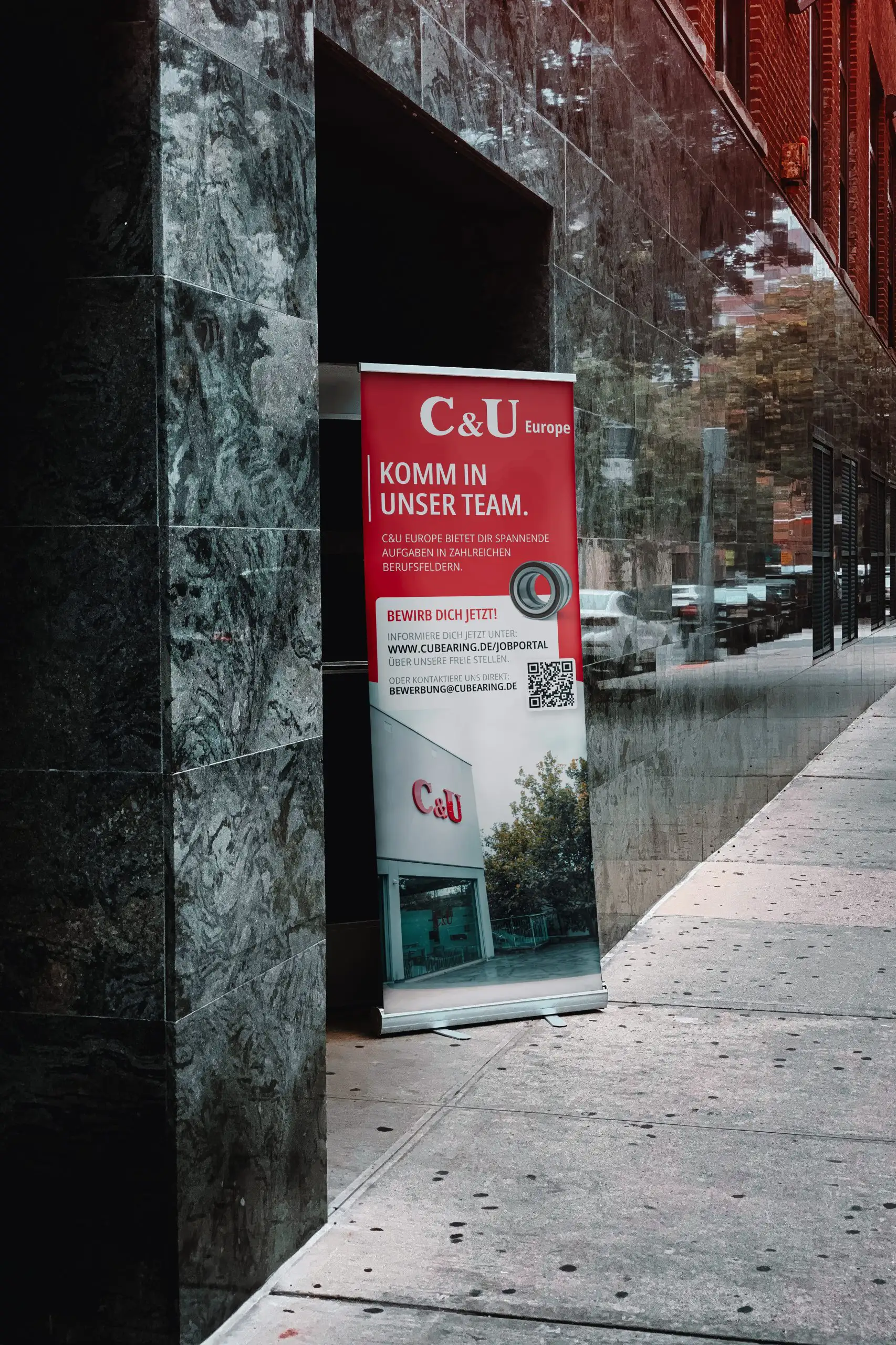 Werbeplakat der Firma C&U Europe vor einem Gebäude aus grau-schwarzen Mamor